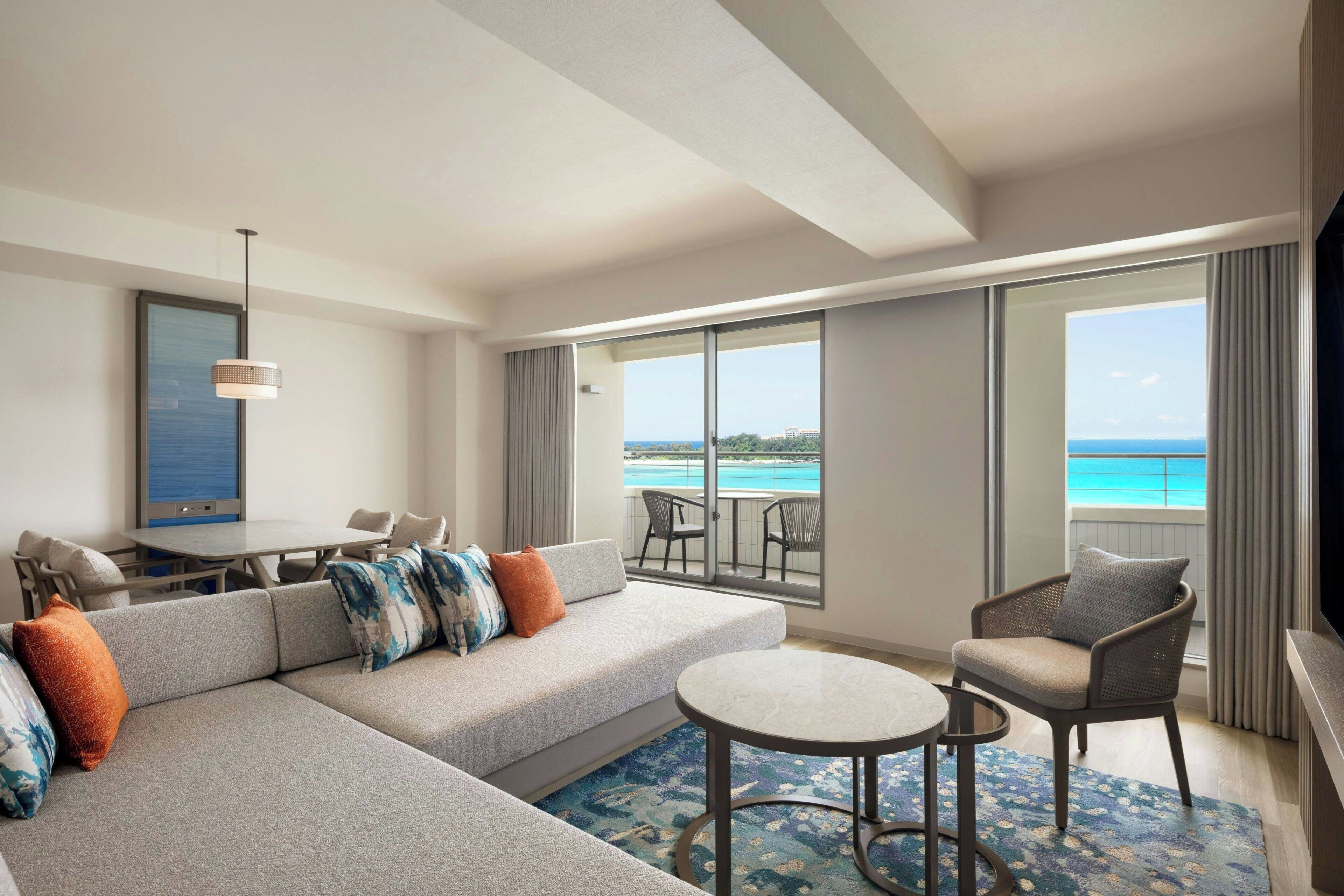 A modern hotel room with a large sofa, a dining table, a small round table, and a balcony with a view of the ocean.