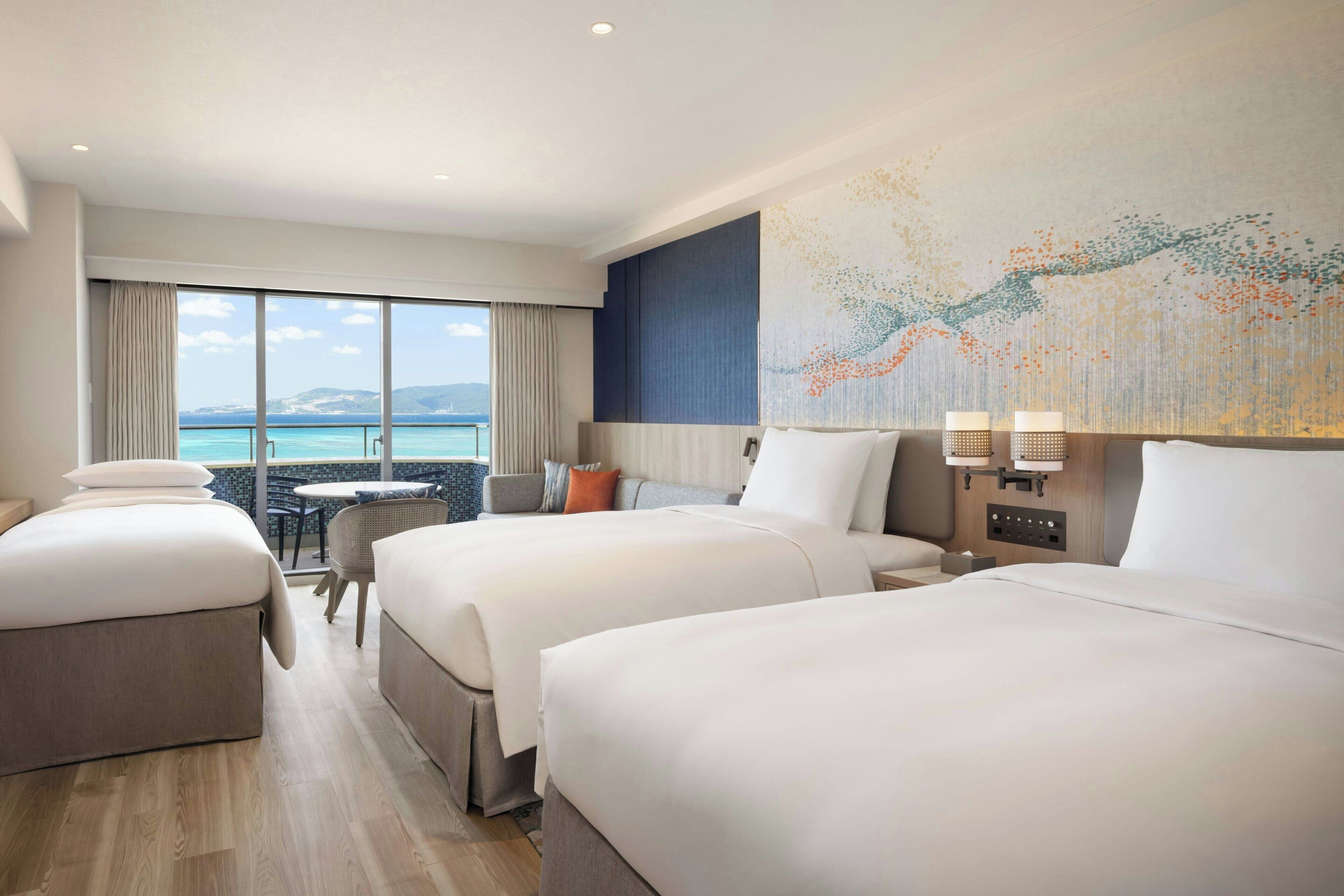 A hotel room with two beds, a dining table, and a view of the sea.
