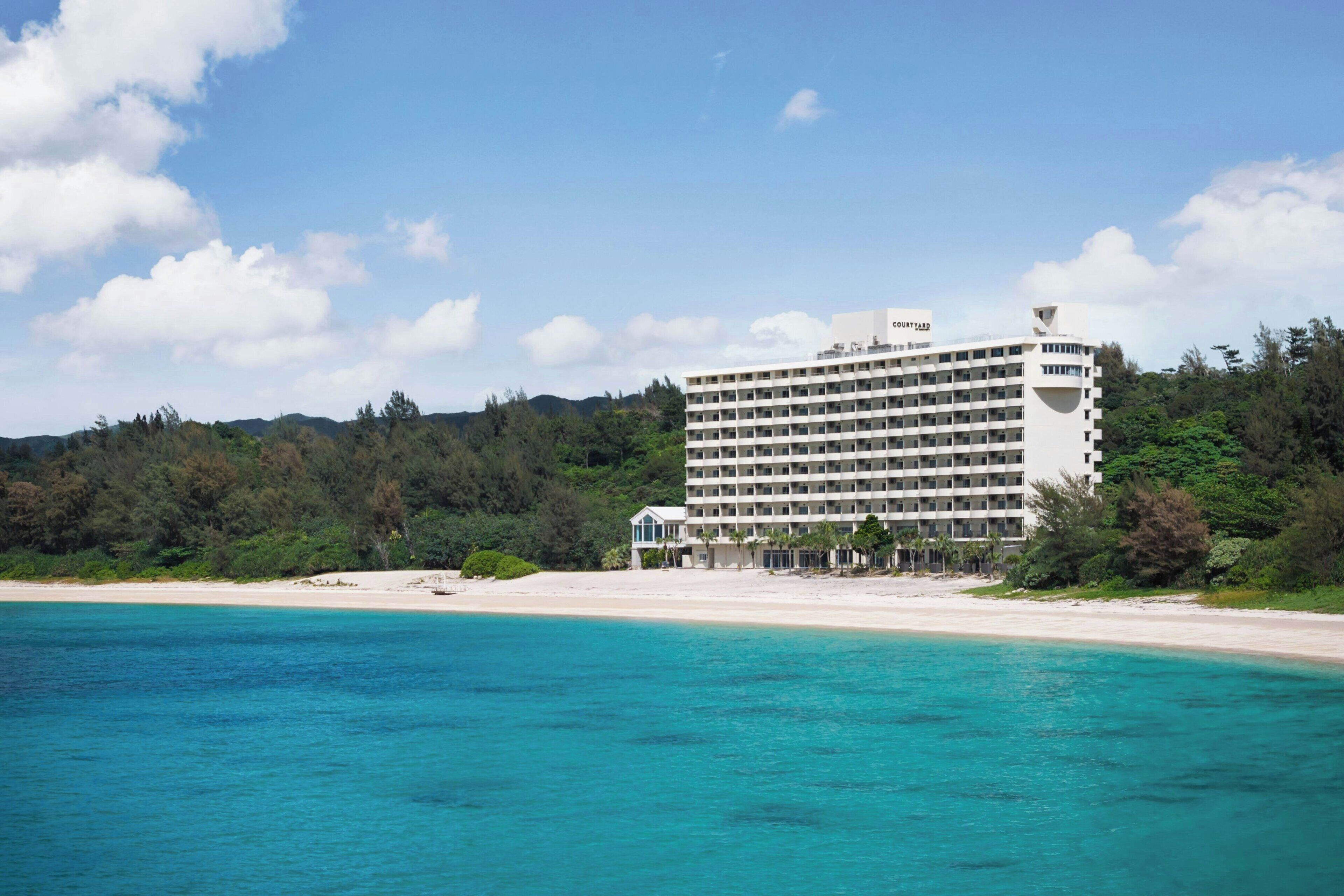 Un hotel grande con varios pisos, una playa al frente y una colina boscosa en el fondo.