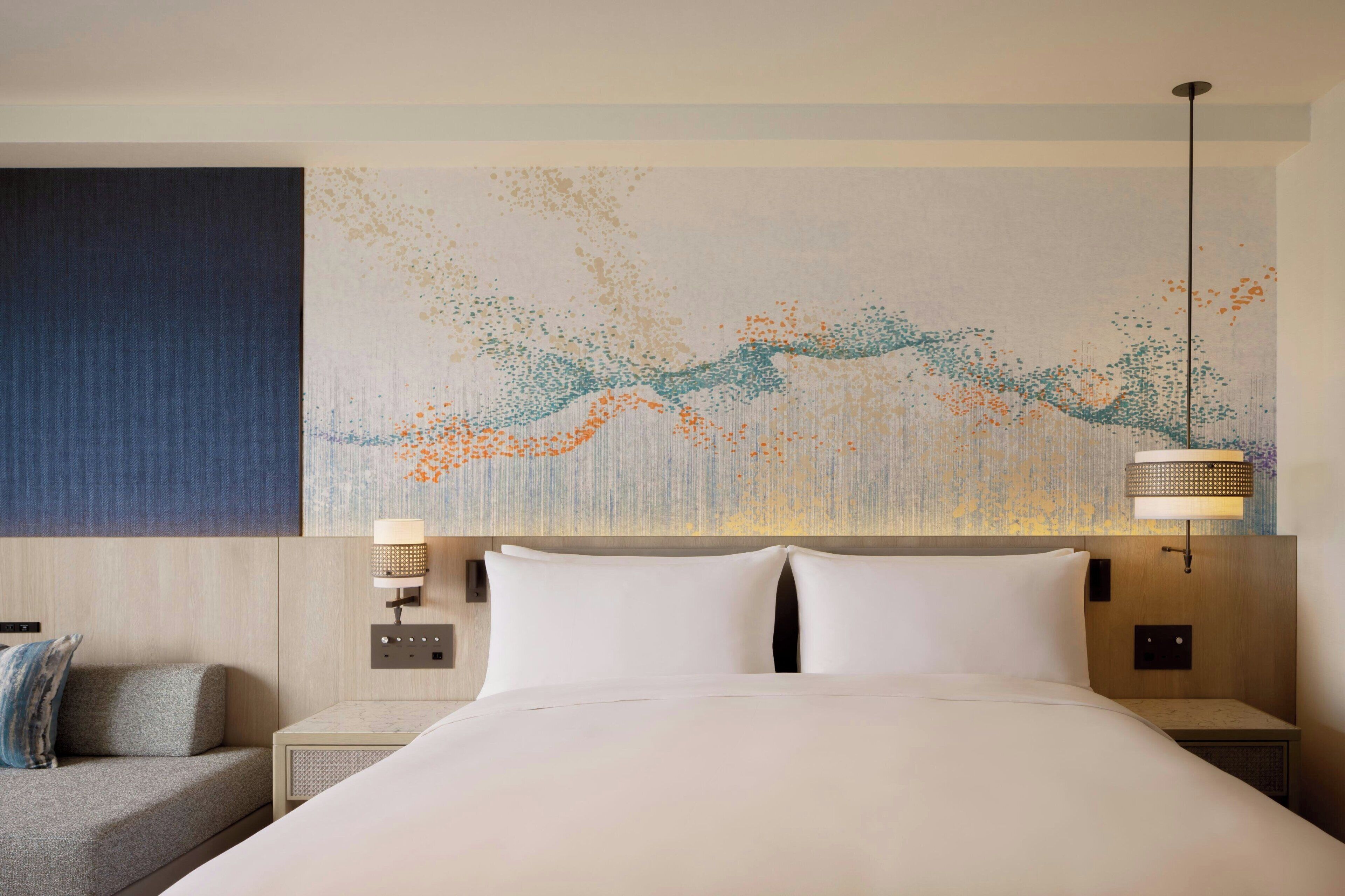A hotel room with a large bed, two bedside tables, a wall art piece, and a nightstand with a lamp.