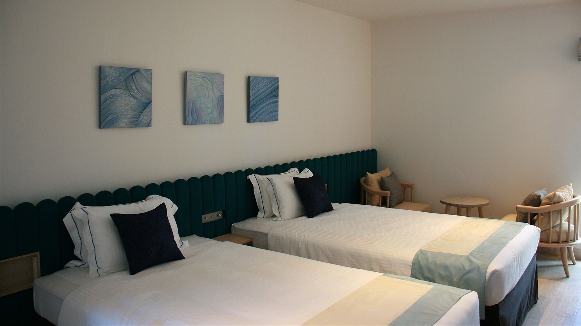 A hotel room with two beds, a small table, a chair, and three wall-mounted paintings.