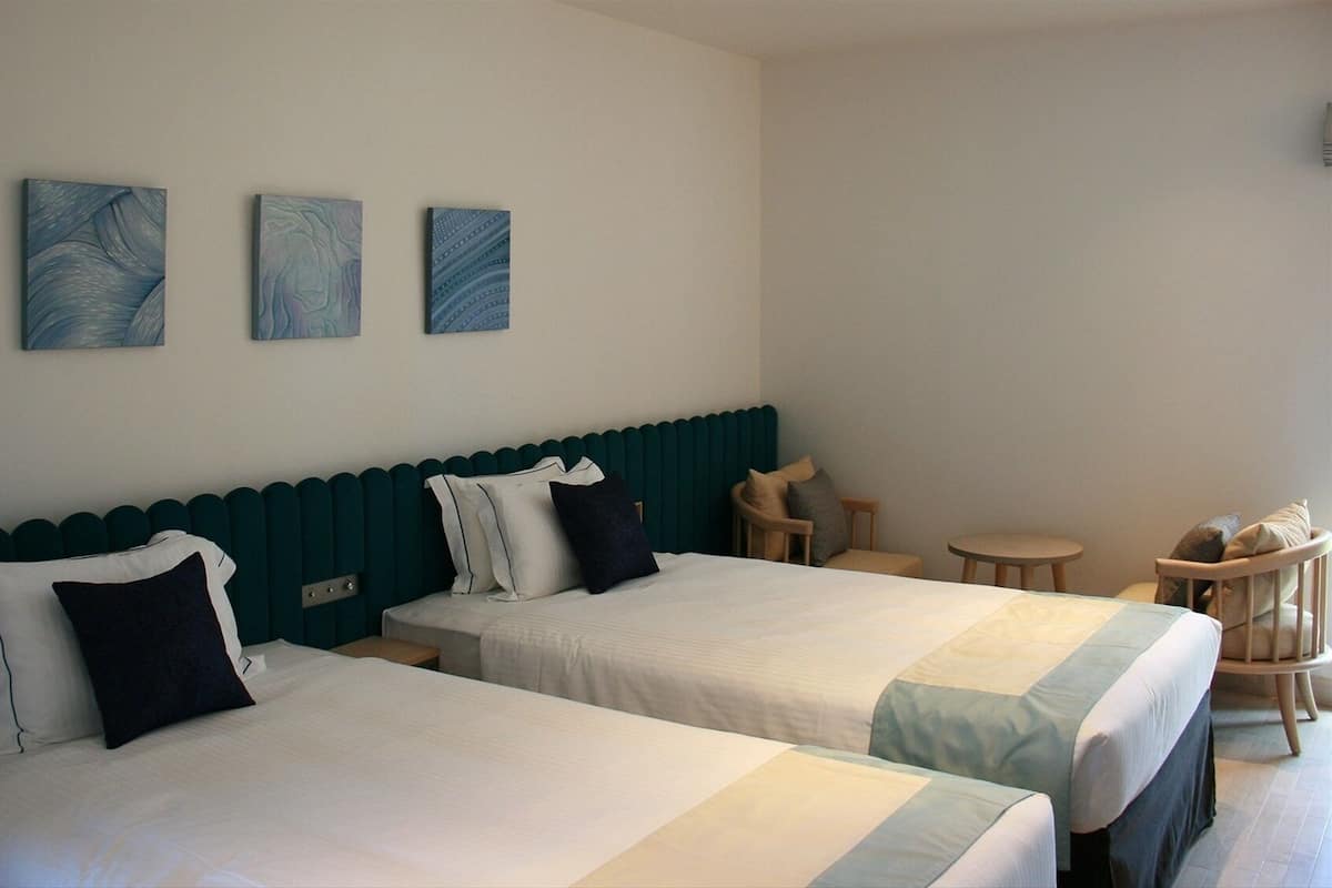 Habitación de hotel con dos camas, una mesita pequeña, una silla y tres cuadros colgados en la pared.