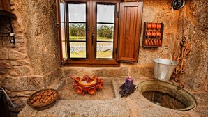 A rustic stone bathroom with a stone sink, a wooden door with a view of the outdoors, a metal bucket, and a basket of nuts. - Casa Castiñeira (Muxía)