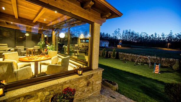 A cozy outdoor seating area with stone walls and a large window overlooking a grassy lawn and a swing set. - Casa Castiñeira (Muxía)