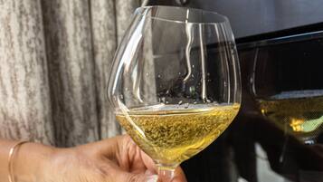 A hand holding a wine glass filled with white wine, with a kitchen and a curtain in the background.