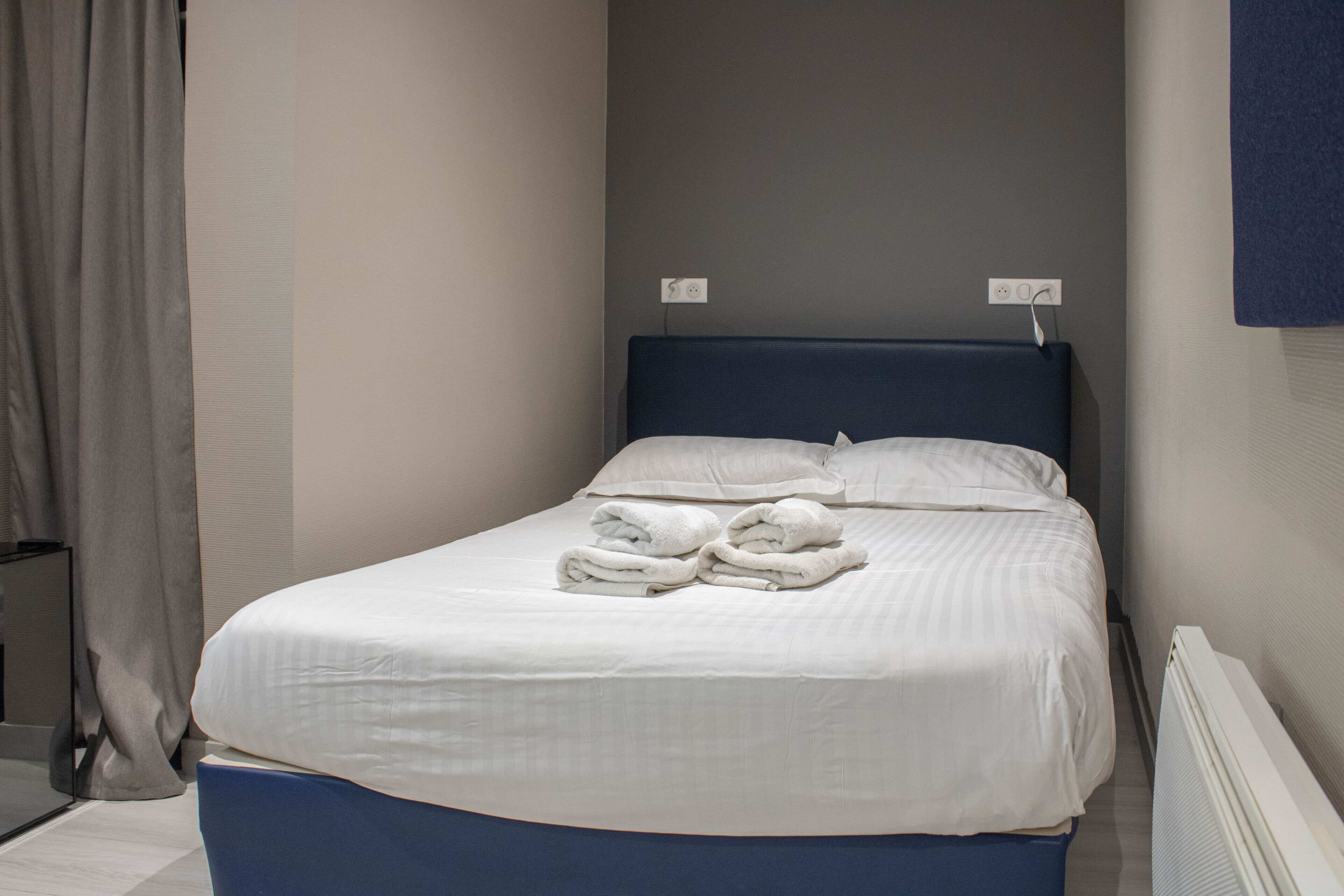 Una cama bien hecha con sábanas blancas y dos toallas dobladas, un cabecero oscuro y una pared lisa con un enchufe.