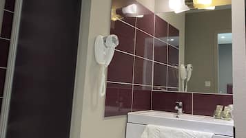 Une salle de bain avec une porte de douche en verre, un lavabo blanc avec une serviette et un miroir dans lequel se reflète un sèche-cheveux.