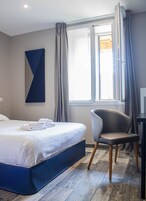 Quarto de hotel moderno com uma cama grande, uma cadeira, uma escrivaninha e uma janela com cortinas.