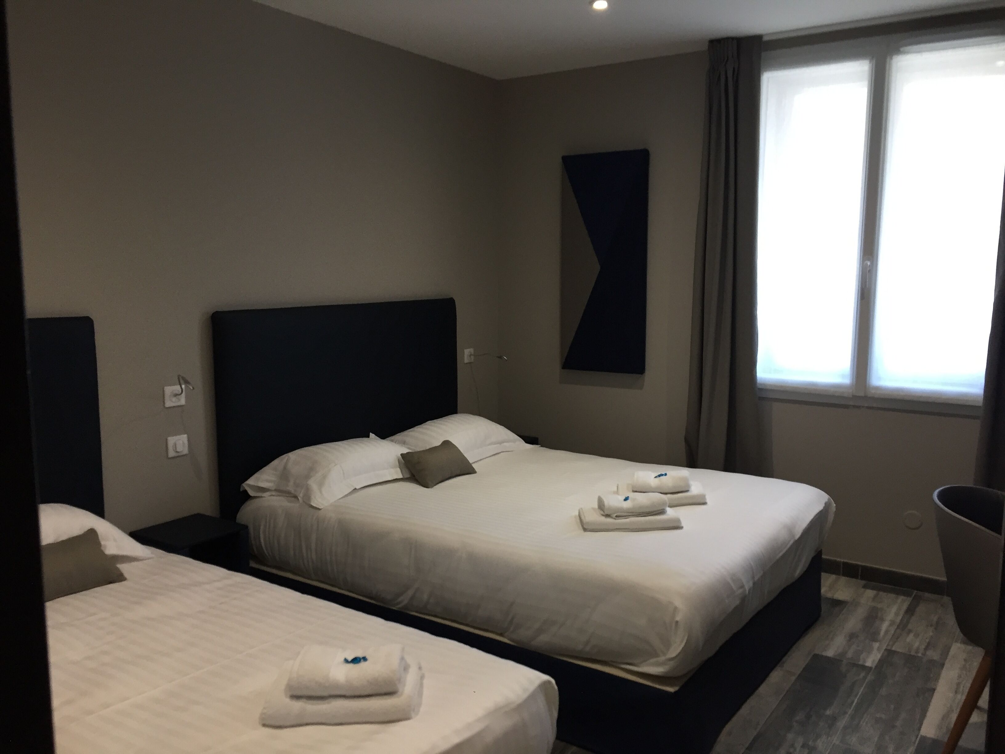 Habitació d'hotel amb dos llits, un capçal de llit fosc, una finestra gran amb cortines i una obra d'art abstracta penjada a la paret.