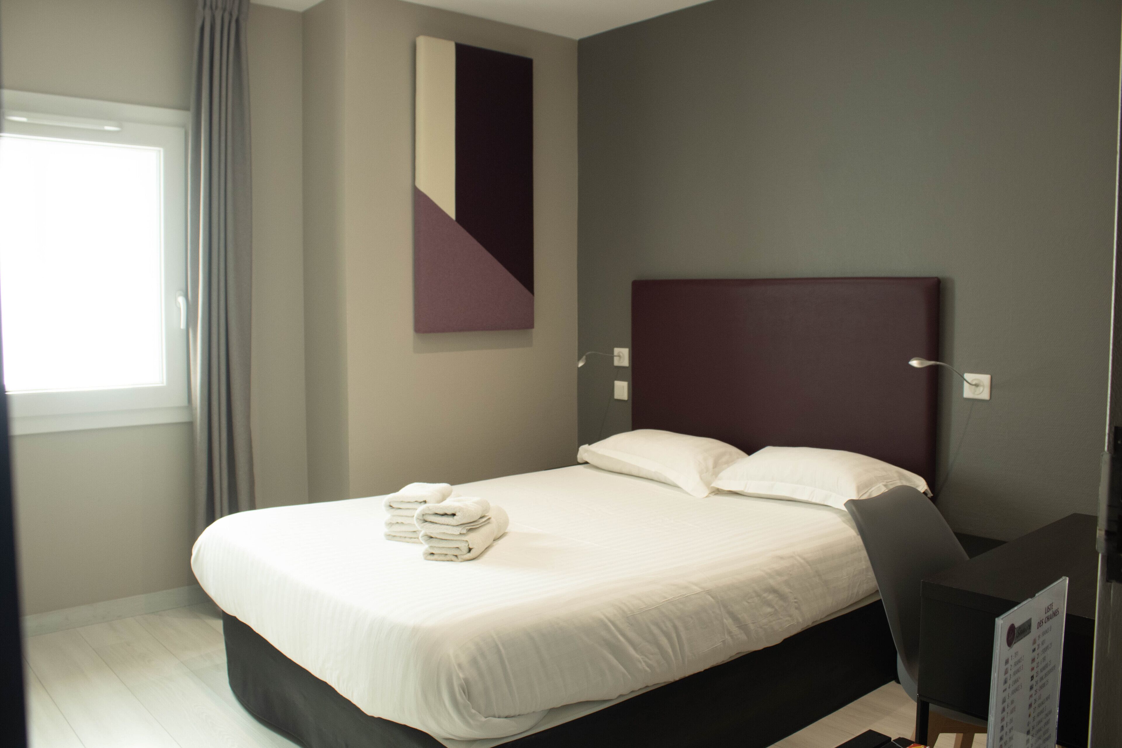 A modern hotel room with a neatly made bed, a large window with curtains, and a wall-mounted artwork.