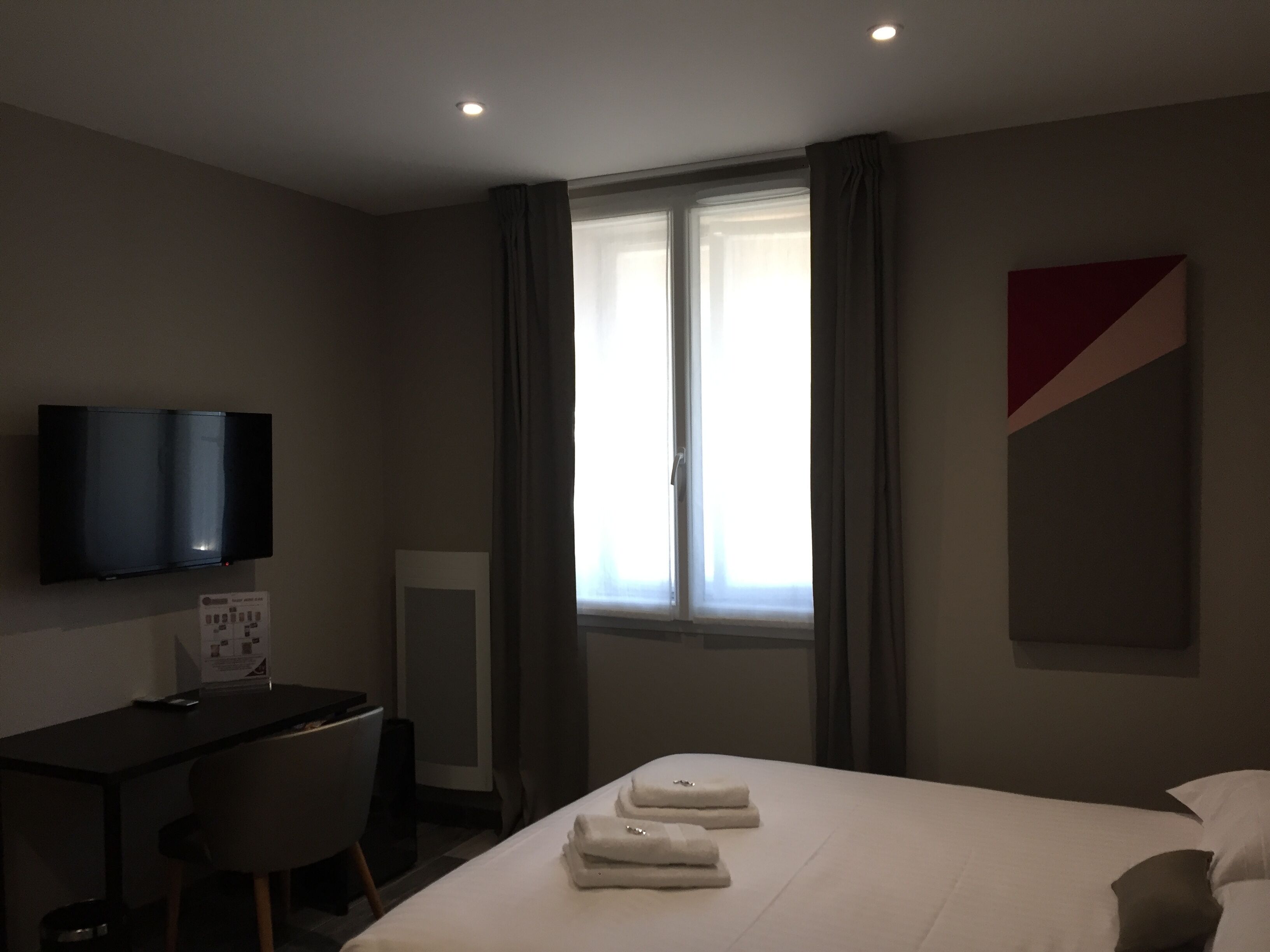 Habitación de hotel con cama, escritorio, silla, televisor montado en la pared y ventana con cortinas.