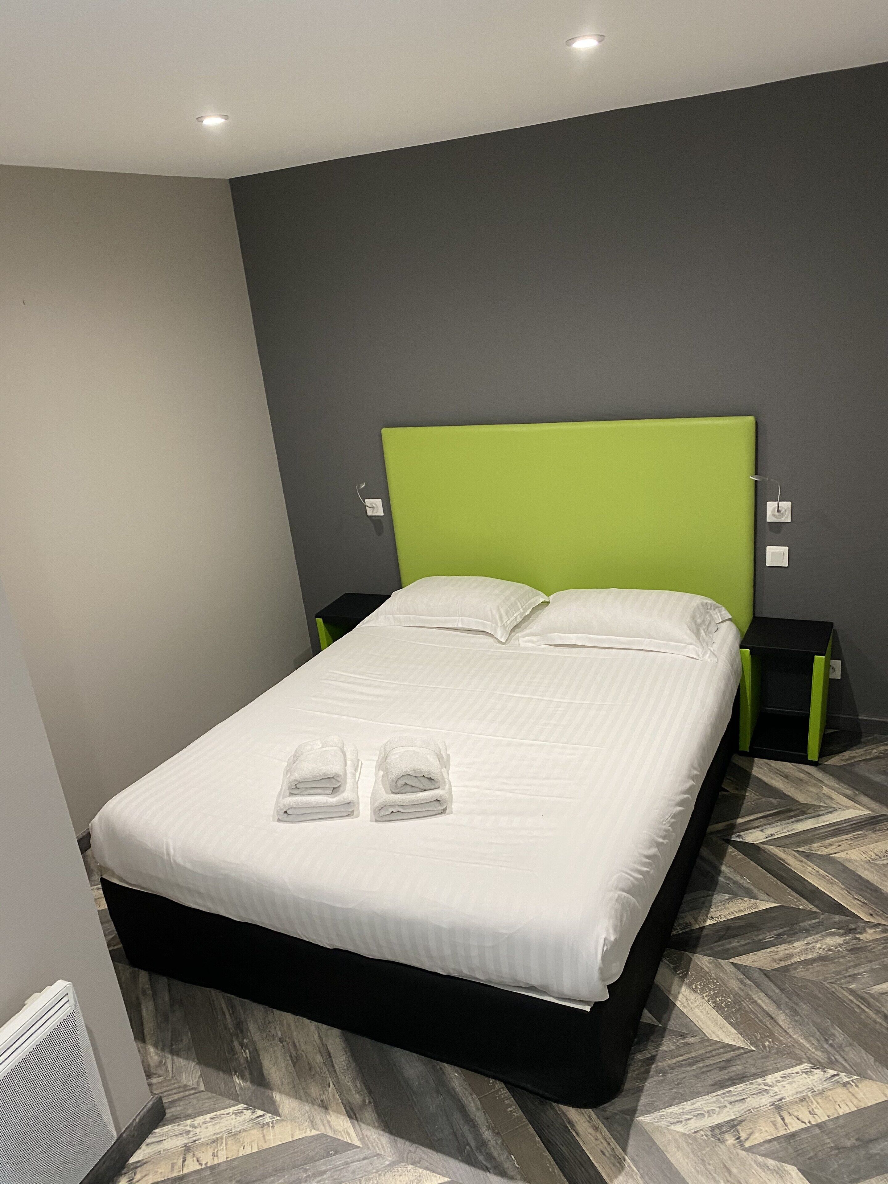 Una cama bien tendida con sábanas blancas y dos toallas, un cabecero verde y una mesita de noche negra con base verde.
