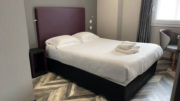 Quarto de hotel com uma cama grande, uma mesinha de cabeceira, uma cadeira e uma janela com cortinas.