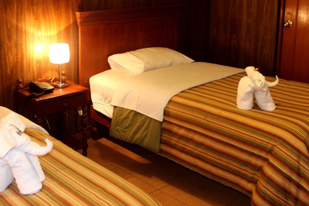 Ein Hotelzimmer mit zwei Betten, jeweils mit einer gestreiften Bettdecke und einem weißen Kopfkissen. An einem Nachttisch stehen eine Lampe und ein Telefon.