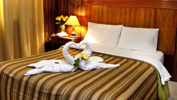 A hotel room with a neatly made bed, a nightstand with a lamp, and a heart-shaped towel arrangement on the bed. - Hotel El Tumi 2 (Huaraz)