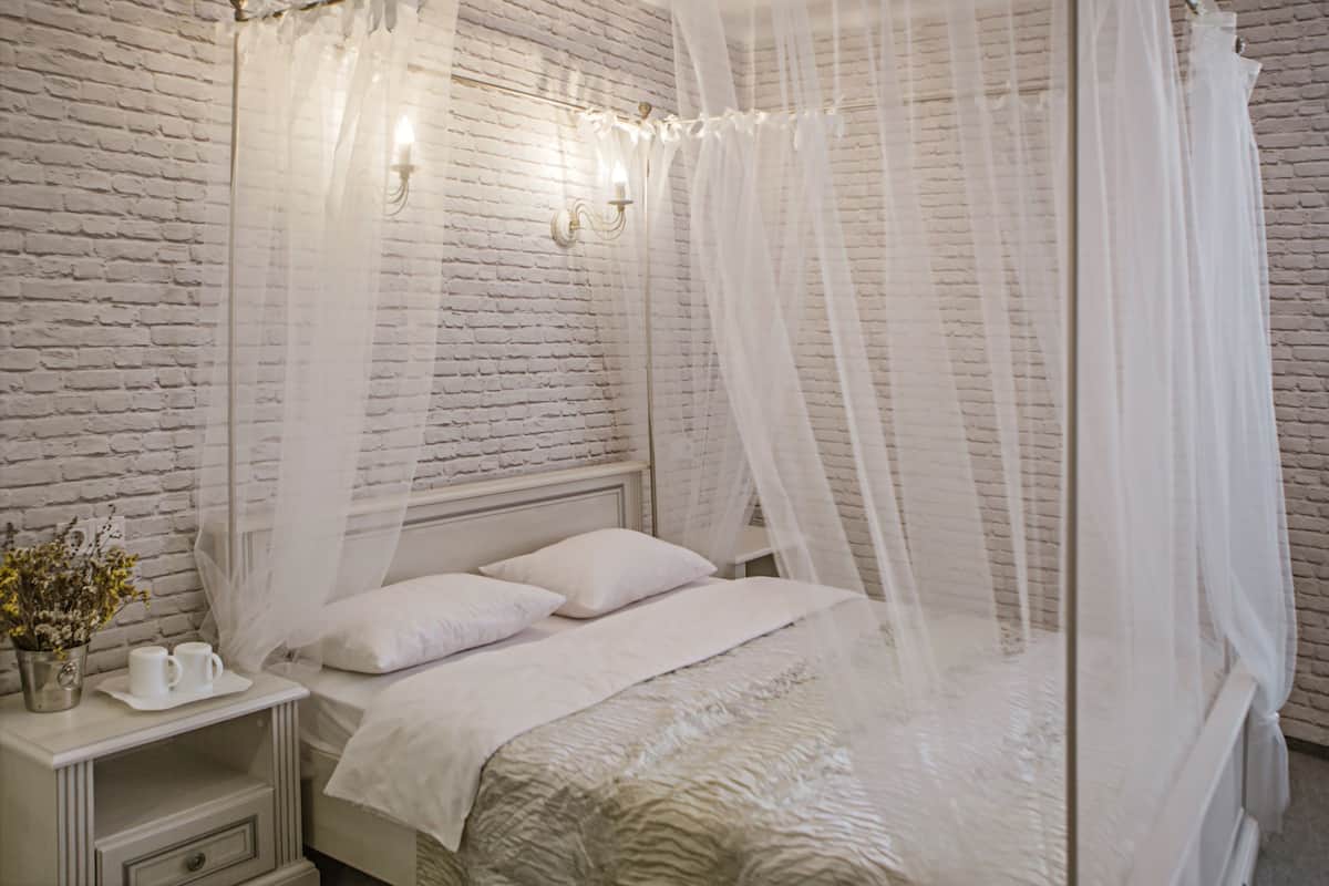 A neatly made bed with a white canopy, a bedside table with a lamp, and a brick-patterned wall.