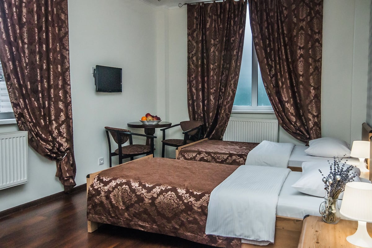 A hotel room with two beds, a desk with a chair, a small table with fruit, and a window with curtains.