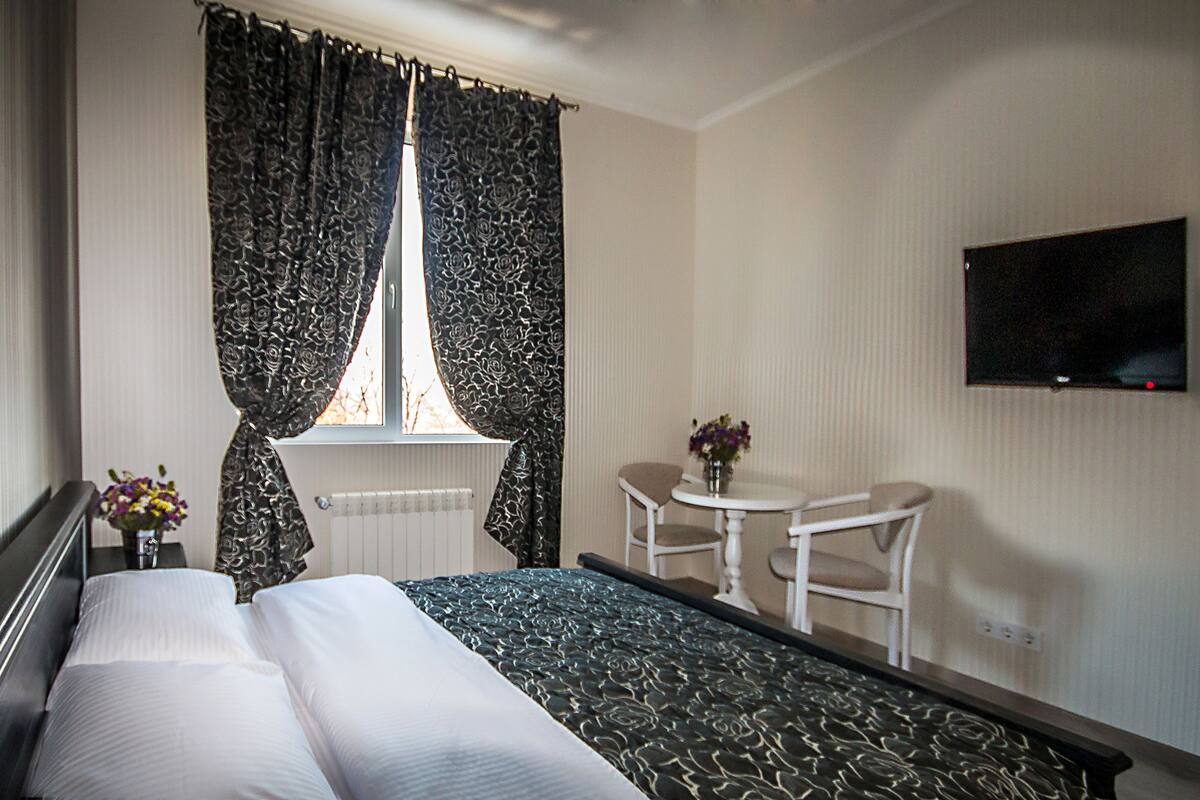 A hotel room with a bed, a window with curtains, a table with a chair, a TV on the wall, and a vase with flowers on the nightstand.