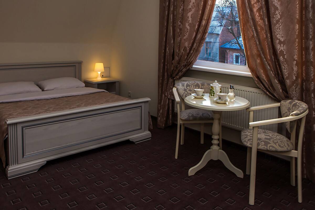 A bedroom with a bed, bedside lamps, a table with a chair, and a view of a building through a window.