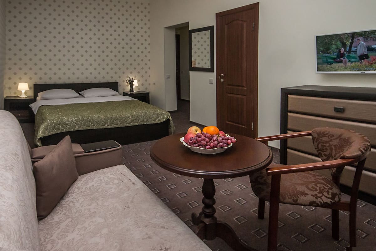A hotel room with a bed, a sofa, a small table with fruit, a chair, a dresser, and a TV on the wall.