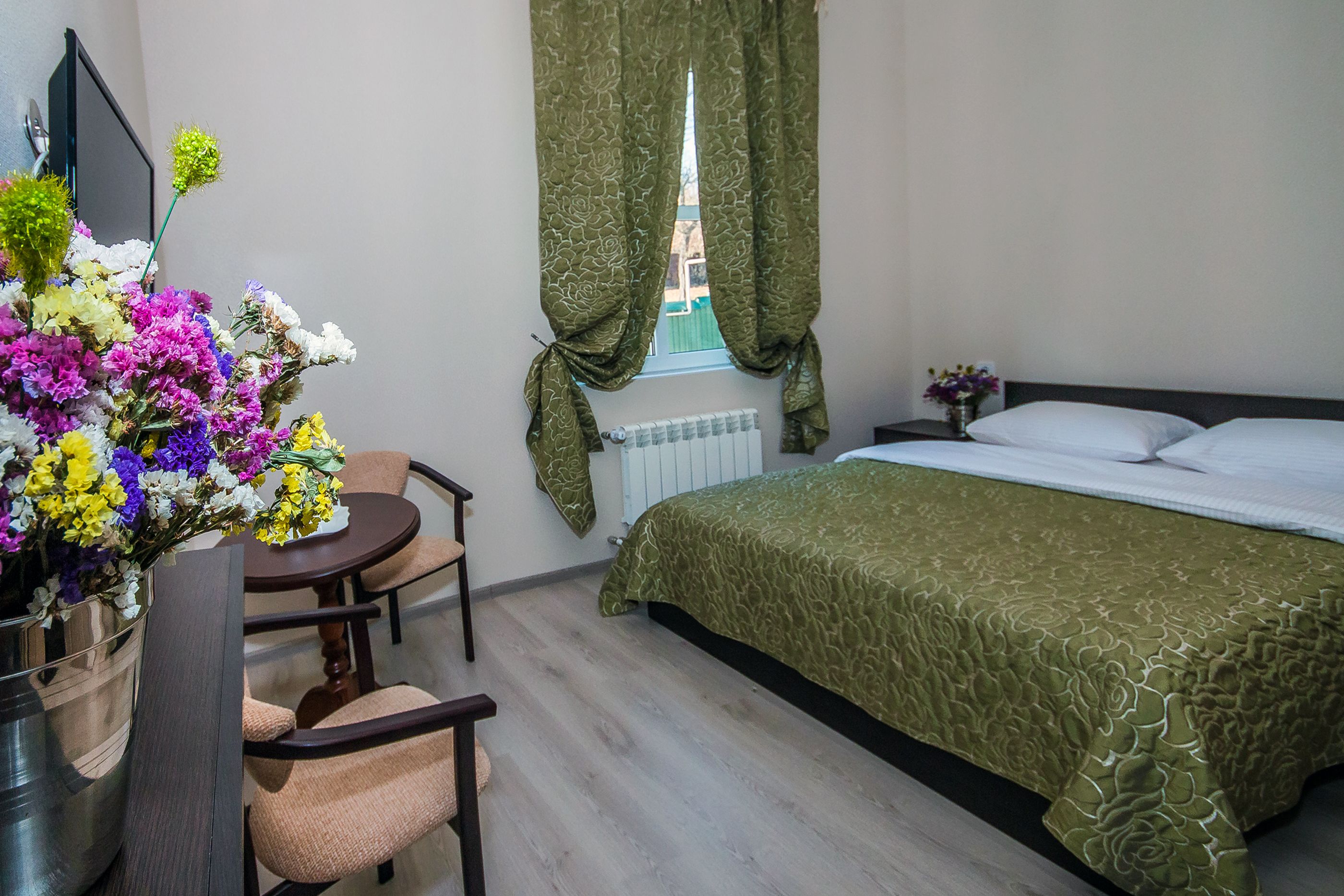 A hotel room with a bed, a desk with a chair, a vase of flowers, and a window with curtains.
