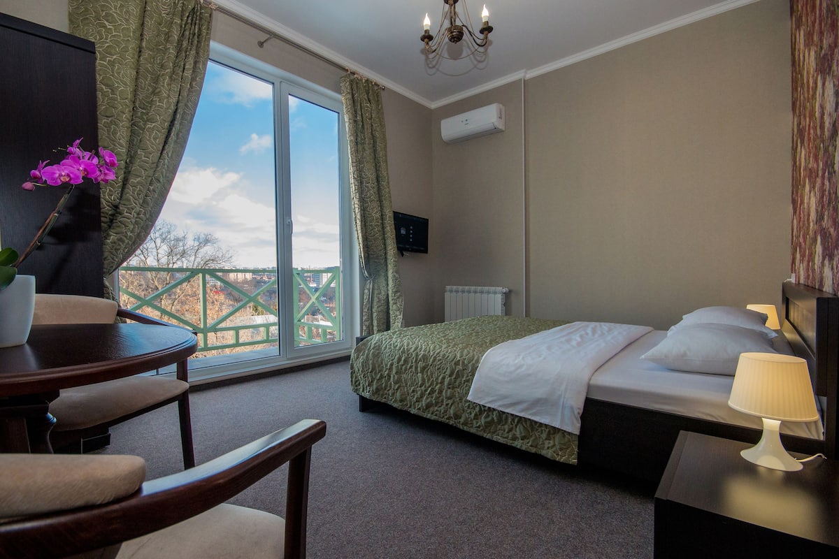 A hotel room with a bed, a desk with a chair, a window with curtains, a chandelier, and a wall-mounted TV.