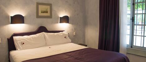 Una cama bien tendida con una colcha a rayas y dos almohadas, un cuadro en la pared y una ventana con cortinas.