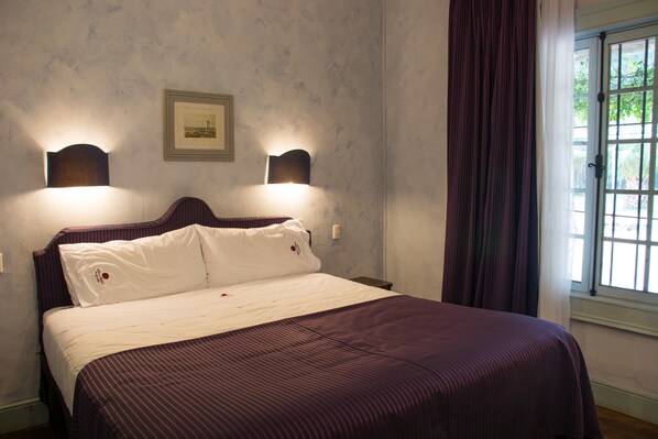 Een netjes opgemaakt bed met een gestreept laken en twee kussens, een ingelijste foto aan de muur en een raam met gordijnen.