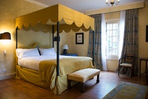 Een hemelbed met een bankje, nachtkastje, stoel en een raam met gordijnen.