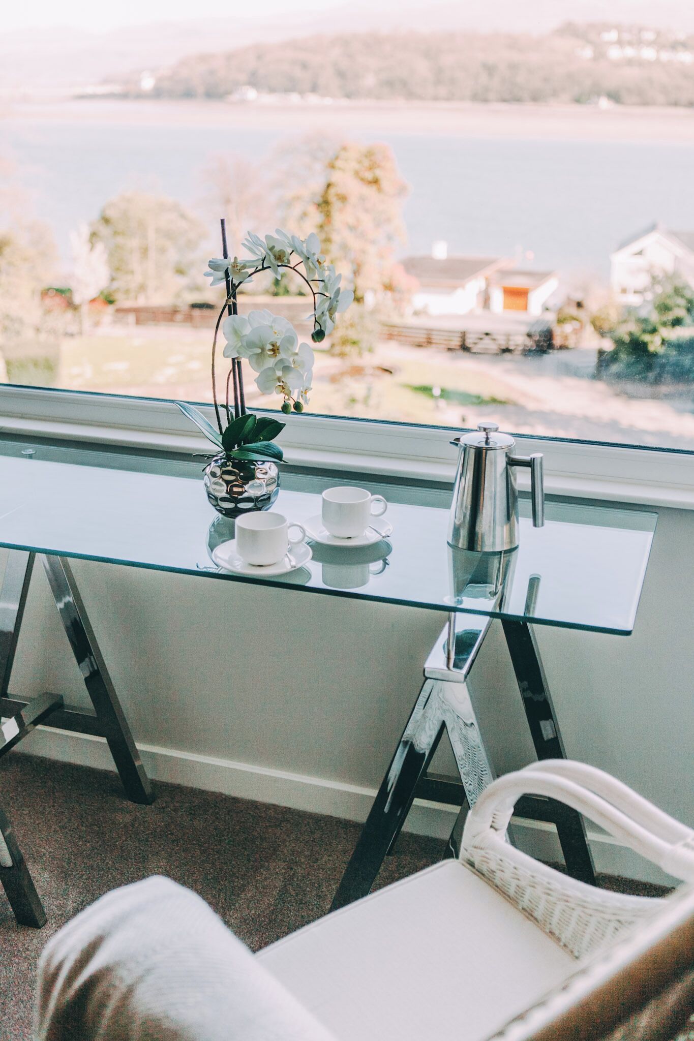 Una mesa de vidrio con dos tazas blancas y una cafetera plateada, una orquídea en maceta pequeña y una silla con cojín blanco.
