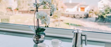 Una mesa de vidrio con dos tazas blancas y una cafetera plateada, una orquídea en maceta pequeña y una silla con cojín blanco.