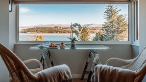 Una habitación con un ventanal grande que da a un paisaje pintoresco, dos sillones de mimbre, una mesita con una taza, un plato de comida y una planta decorativa.