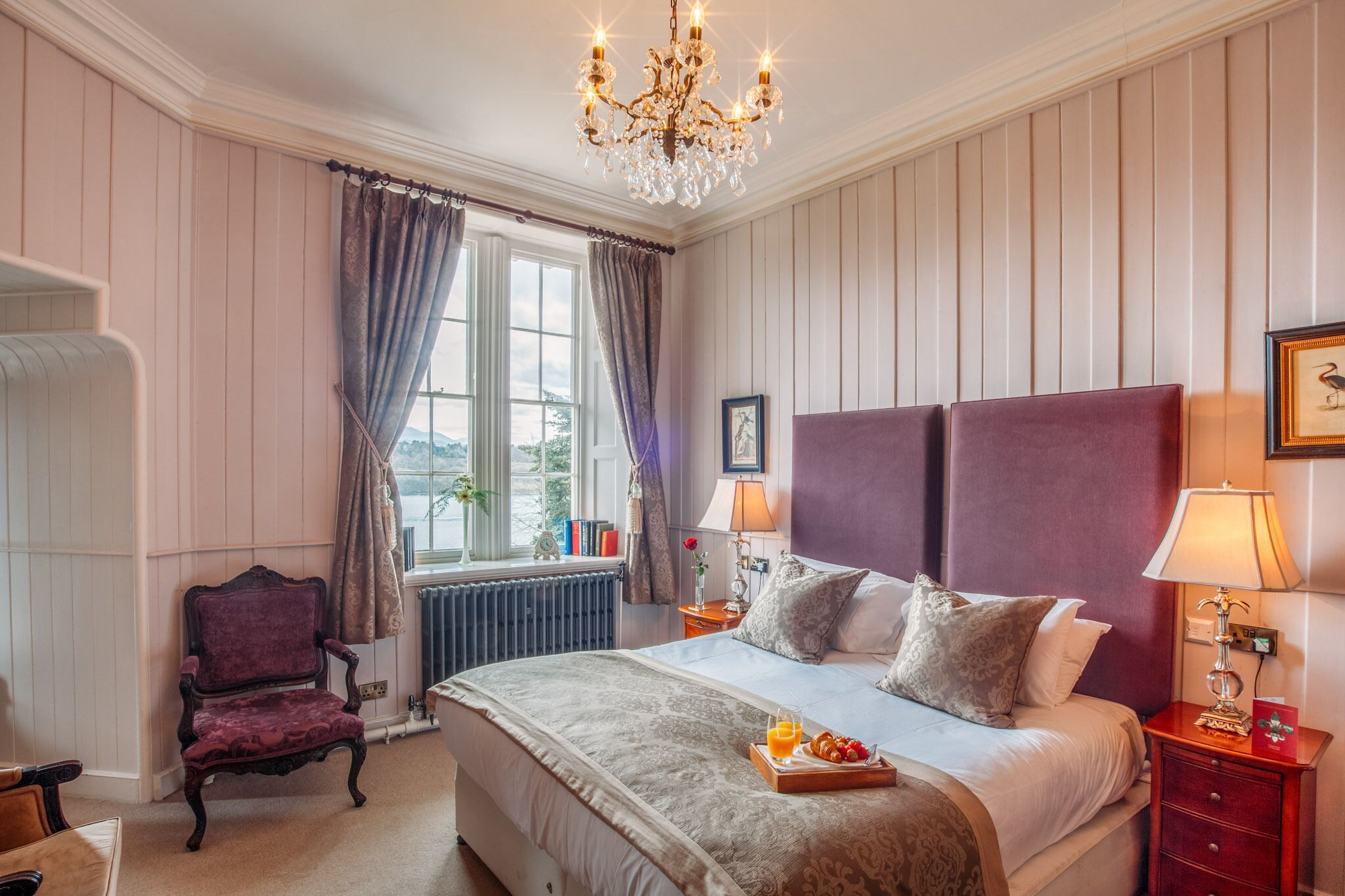 Ein Hotelzimmer mit einem grossen Bett, einem Chandelier, einem fenster mit Vorhängen, einem Sessel und einem Nachttisch mit Lampe.