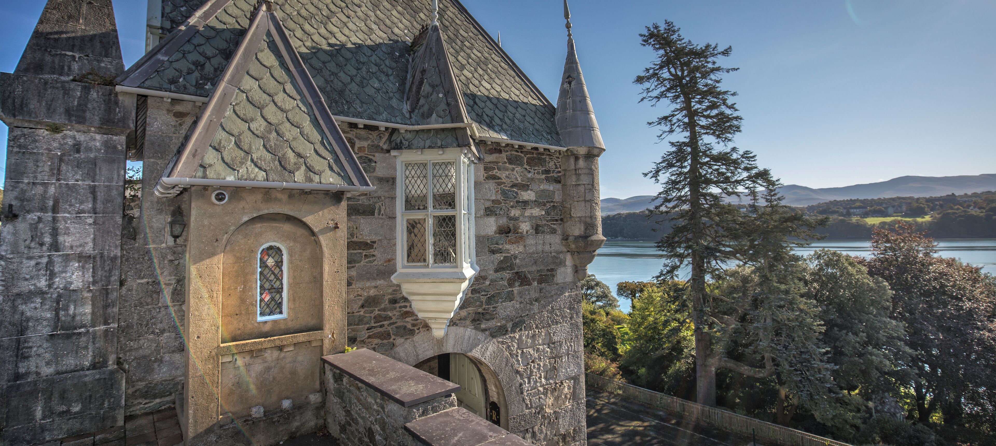 Un edificio de piedra con un techo empinado, una ventana con celosía decorativa y vistas a un lago y un bosque.
