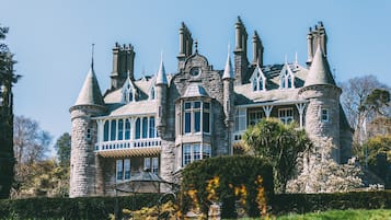 A large, historic stone building with multiple towers and turrets, surrounded by lush greenery and a well-maintained garden.