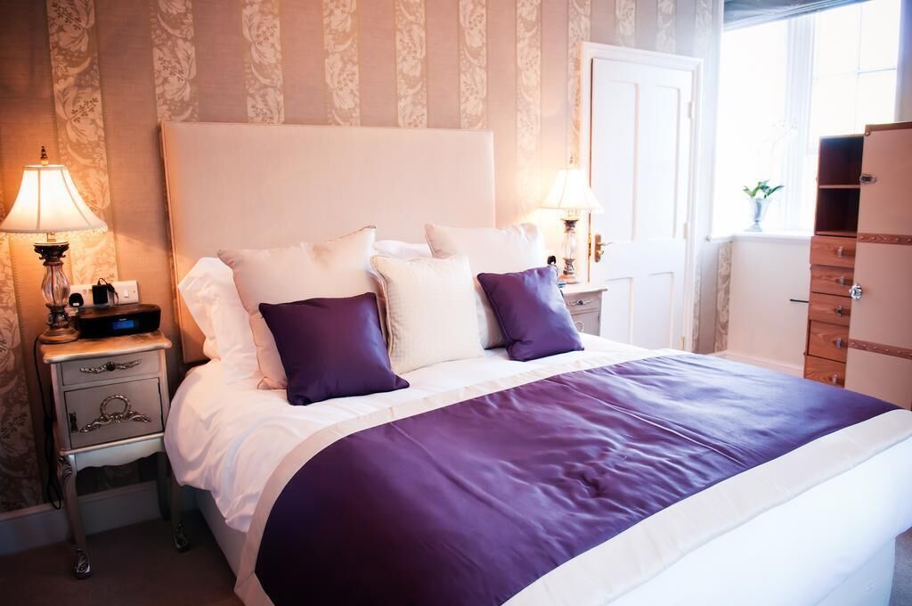 Una cama bien hecha con cabecera blanca, dos almohadas moradas y colcha blanca y morada. Hay dos lámparas de noche a ambos lados de la cama y un aparador de madera a la derecha.