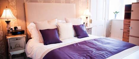 Una cama bien hecha con cabecera blanca, dos almohadas moradas y colcha blanca y morada. Hay dos lámparas de noche a ambos lados de la cama y un aparador de madera a la derecha.