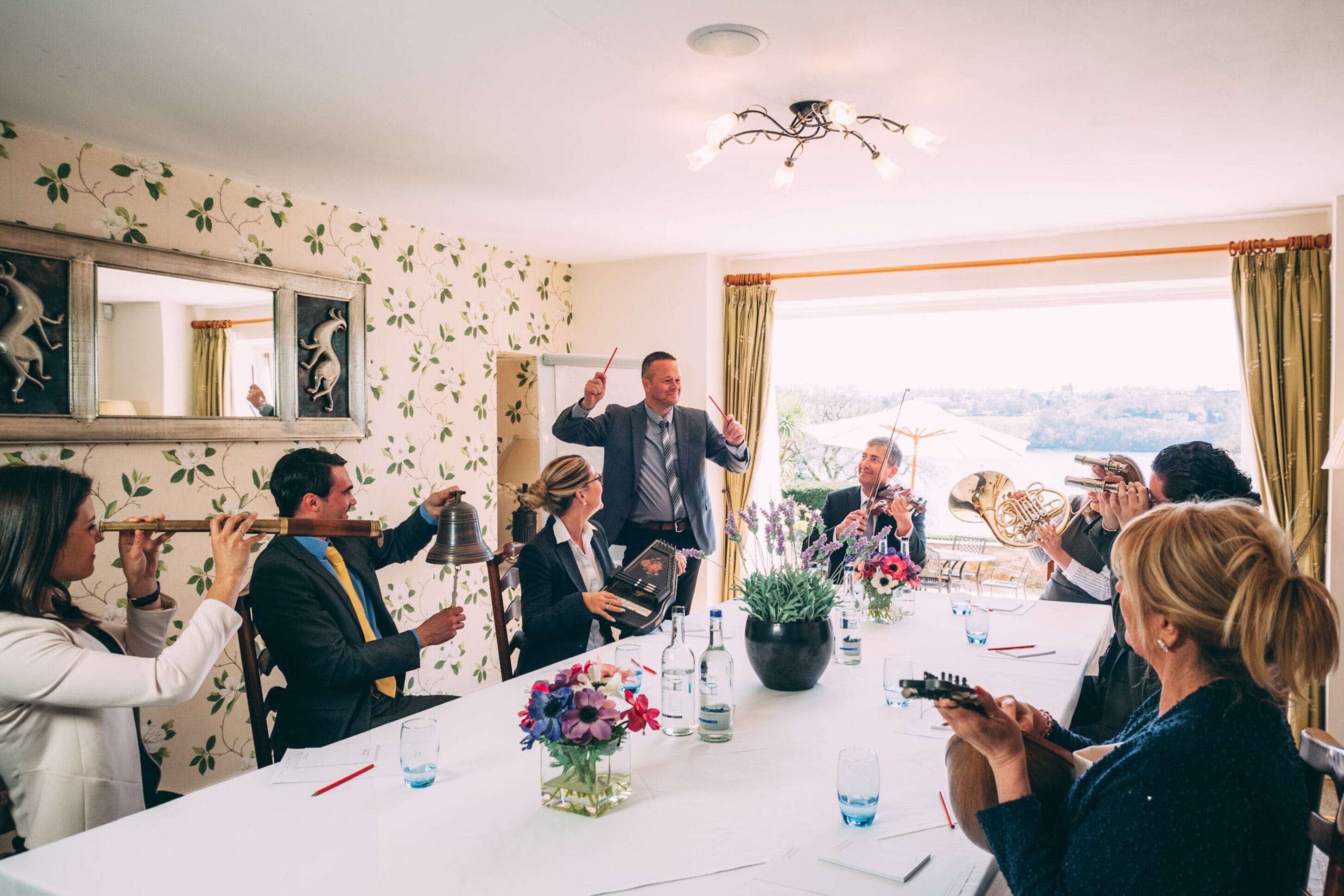 A group of people seated around a table in a room with floral wallpaper, a chandelier, and a view through large windows.