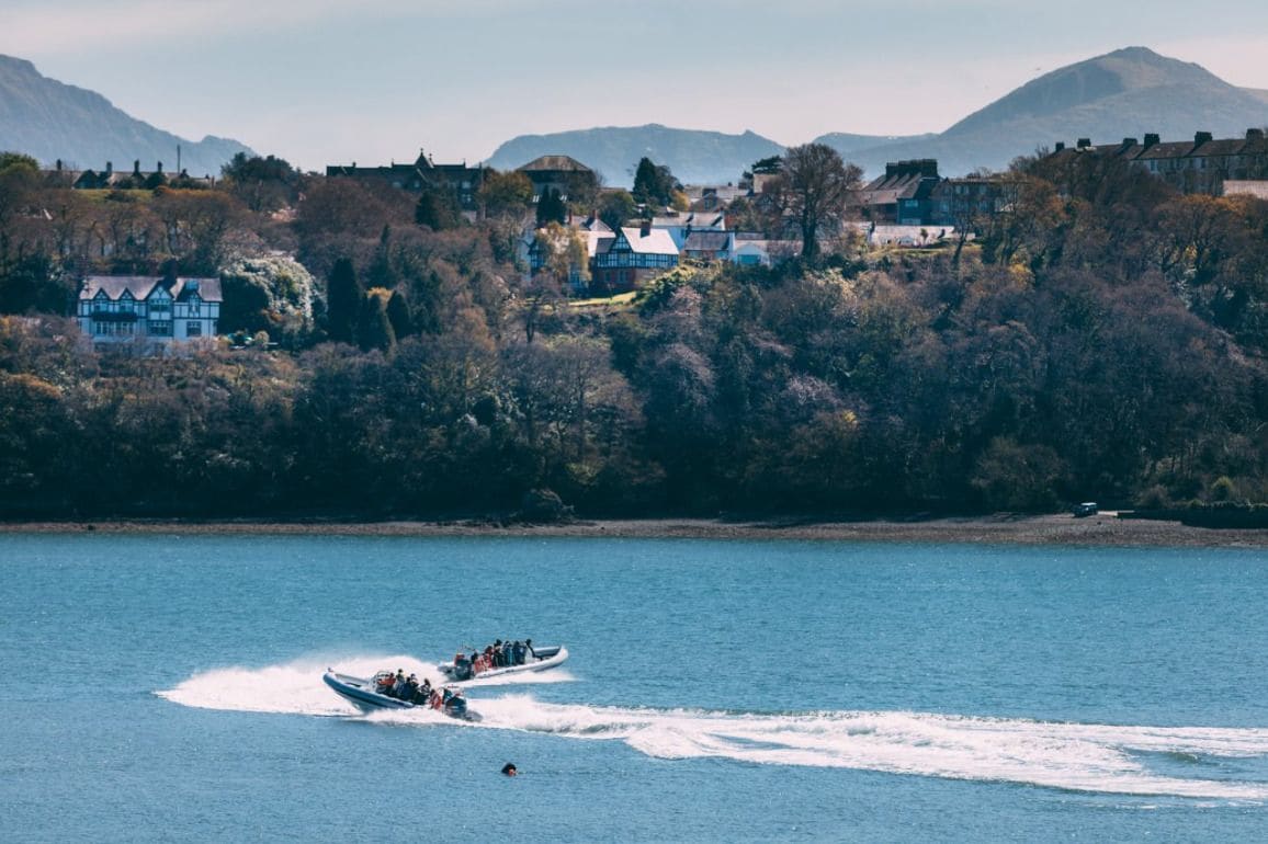 Un vaixell surant ràpidament sobre un llac tranquil, amb una zona residencial i muntanyes al fons.