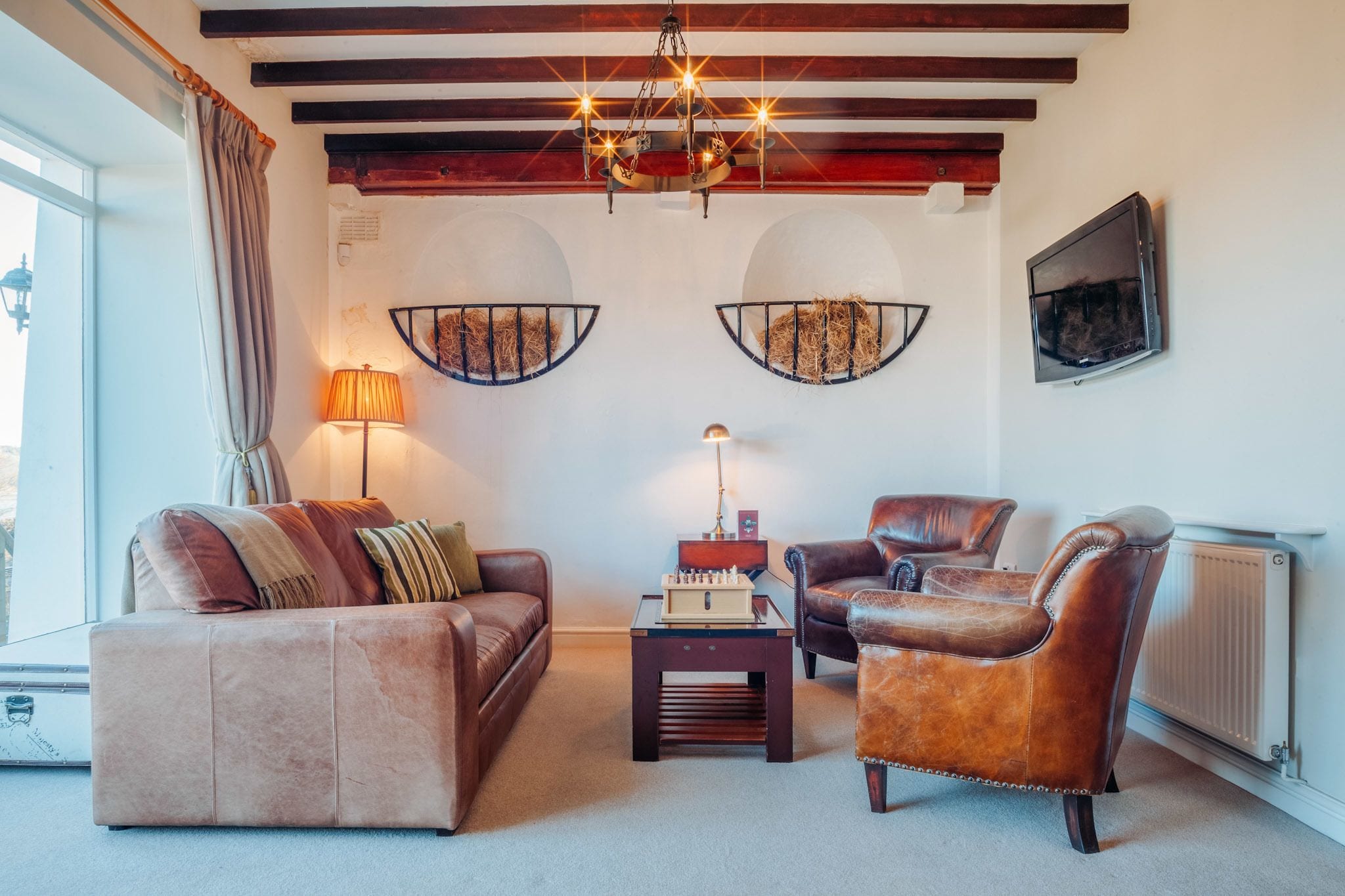 Een knusse woonkamer met een bruine leren bank, een lederen fauteuil en een houten salontafel met een schaakbord. Een kroonluchter hangt aan het plafond, en er zijn twee wandplanken met hooibalen.