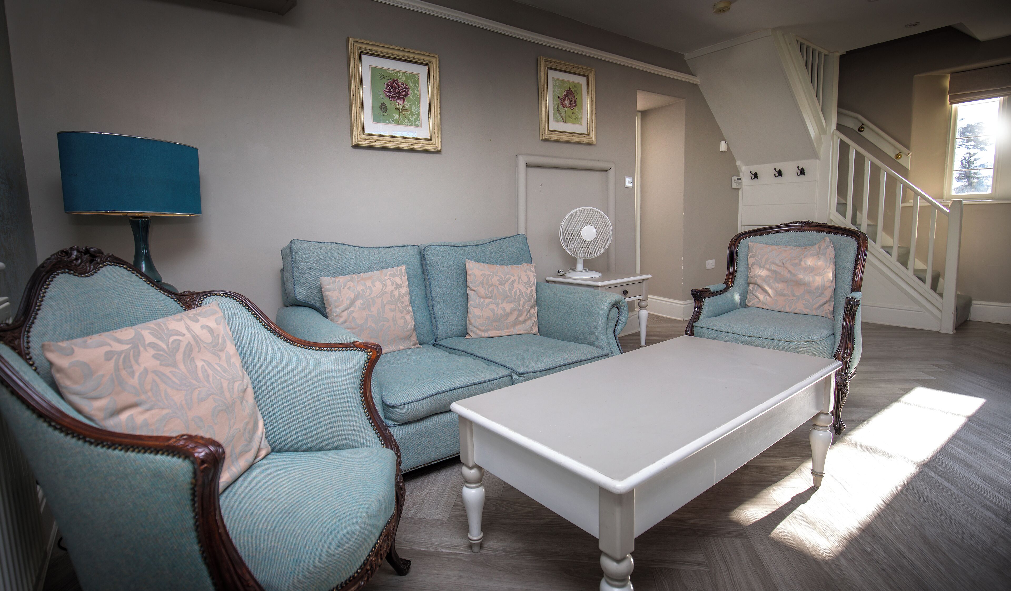 A living room with a blue sofa, matching armchairs, a white coffee table, and a fan. There are two framed botanical prints on the wall.