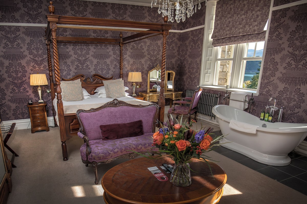 Una cama con dosel sostenido por cuatro columnas, con estructura de madera, una butaca morada tapizada, una mesa redonda de madera con un jarrón de flores y una bañera con patas de garra.