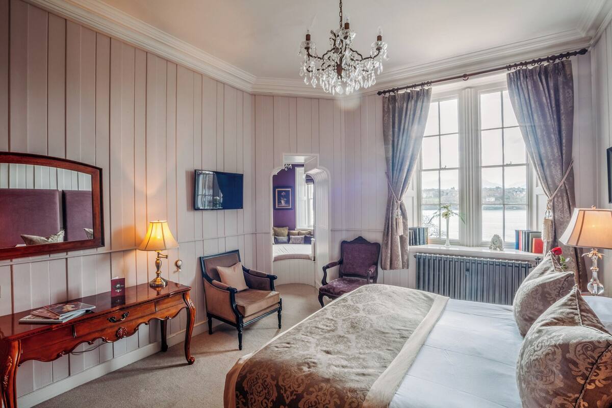Habitación de hotel con una cama grande, dos sillones, una mesa consola de madera, un espejo, un televisor y vistas al exterior a través de una ventana.