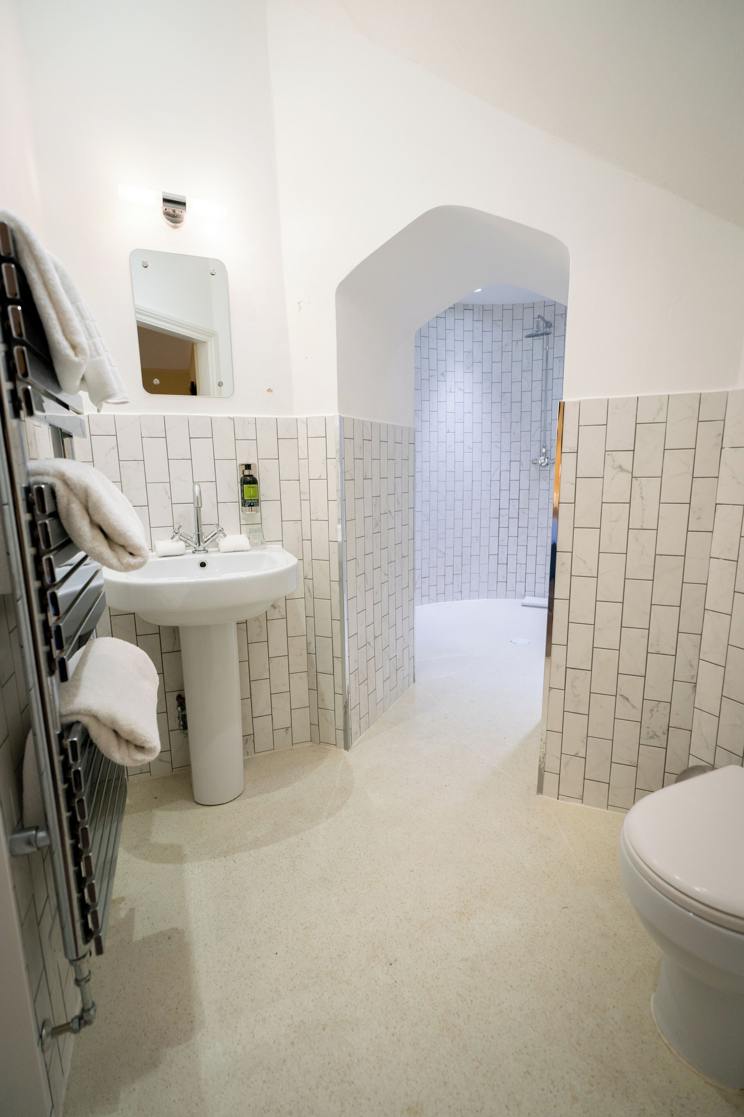 A bathroom with a white sink, a mirror, and a toilet. There is a towel rack with towels and a bottle of hand soap on the sink. The walls are tiled, and there is a shower visible in the background.