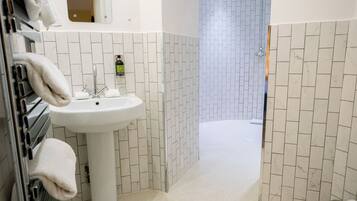 A bathroom with a white sink, a mirror, and a toilet. There is a towel rack with towels and a bottle of hand soap on the sink. The walls are tiled, and there is a shower visible in the background.