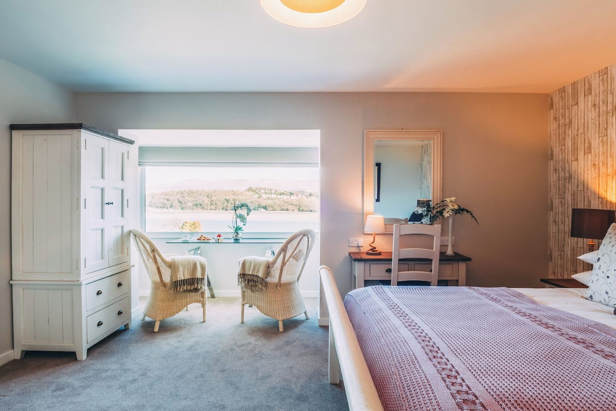 Un dormitorio con una cama grande, un aparador, dos sillas, un escritorio y vistas al exterior.