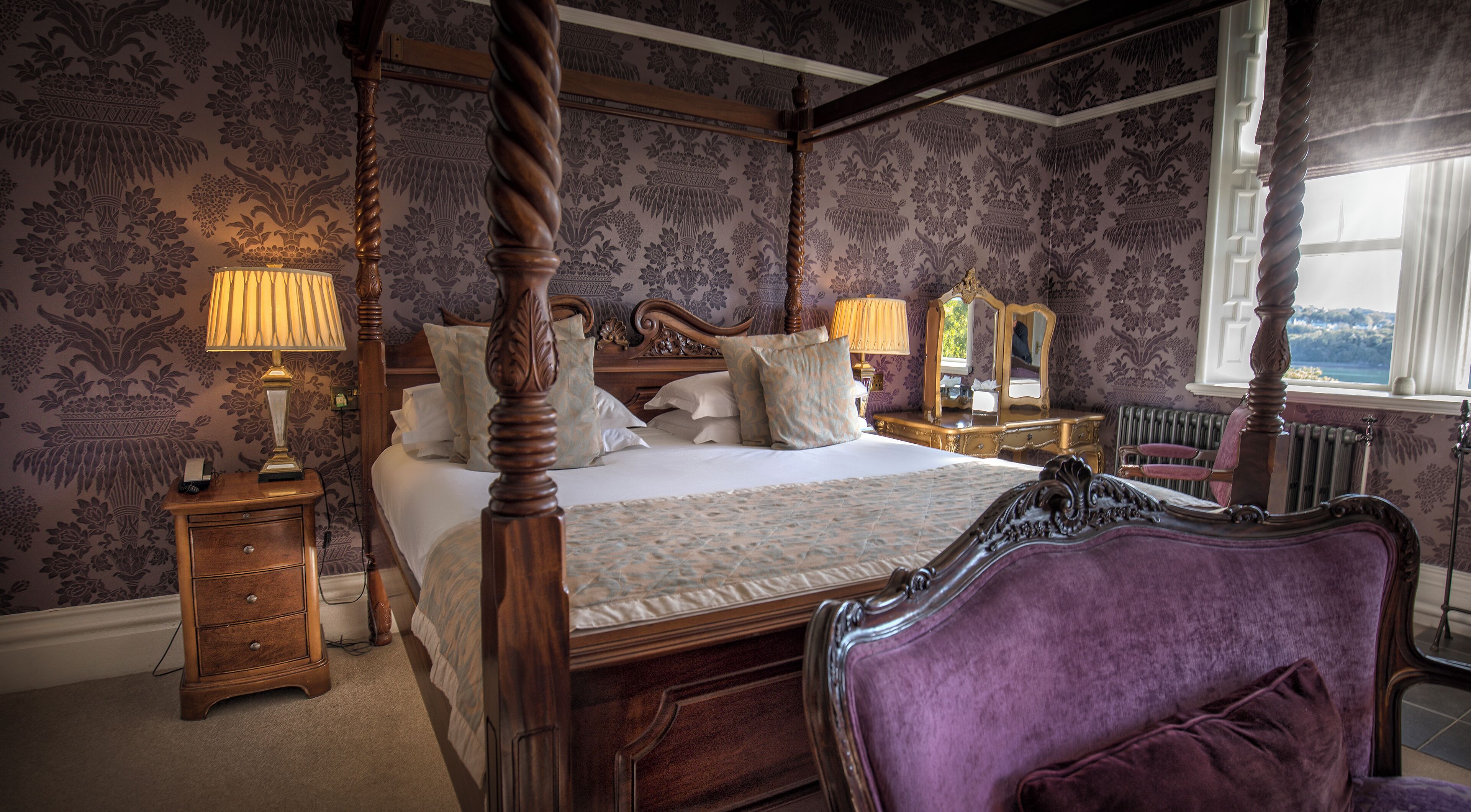 A four-poster bed with a canopy, a wooden nightstand with a lamp, a dresser, and a purple armchair.