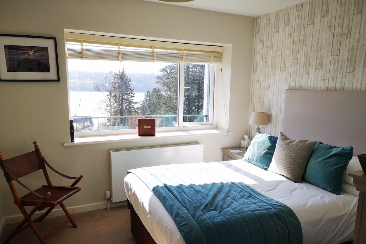 Un dormitorio con cama, ventana con vistas, una silla y una mesita de noche con lámpara.