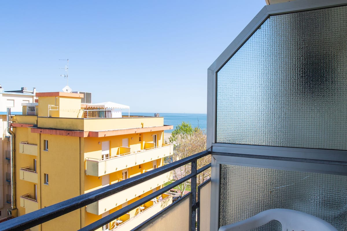 A balcony with a white plastic chair, a view of the sea, and buildings in the background.