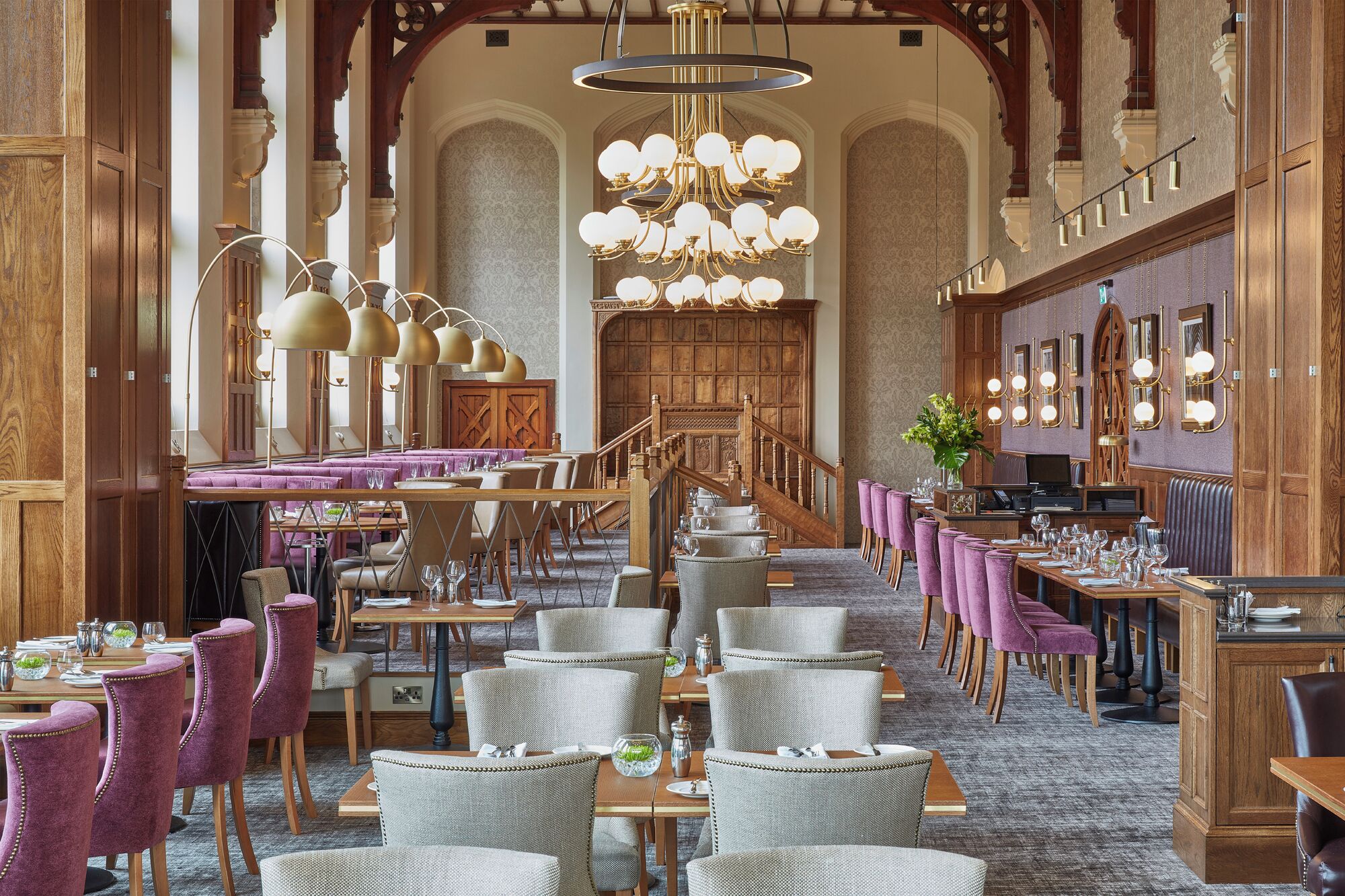 A spacious hotel dining area with multiple tables and chairs, a grand staircase, and ornate chandeliers.